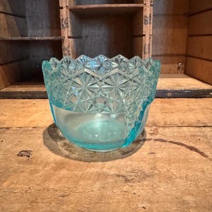 May include: A clear, light blue glass bowl with a diamond pattern around the rim. The bowl sits on a wooden surface, with a wooden crate in the background. The bowl is likely vintage or antique.