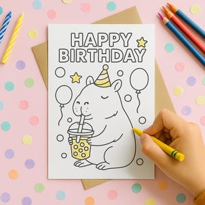 May include: A white birthday card with the words "HAPPY BIRTHDAY" and a cartoon capybara drinking from a bubble tea cup. The card is being colored with a yellow crayon. Colored pencils and confetti are in the background.