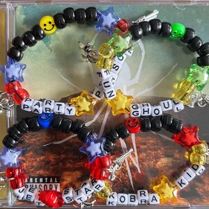 May include: A collection of colorful beaded bracelets with star and smiley face charms. The bracelets spell out "PARTY", "GHOUL", and "KID KOBRA" with white letter beads. The bracelets are arranged on a CD case.