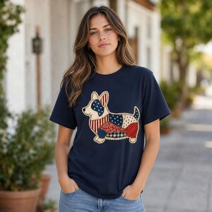 Puede incluir: Una camiseta azul marino de manga corta con un gráfico central de un perro Corgi. La ilustración del perro está compuesta por varios patrones de tela estilo patchwork, incluyendo rayas rojas y blancas, azul con estrellas blancas y motivos de cachemira rojos.