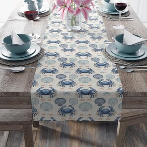 May include: A beige table runner with a repeating pattern of blue crabs, seashells, and small orange fish. The runner is set on a wooden table with place settings, including blue bowls, plates, and wine glasses. A vase of pink flowers sits in the center.