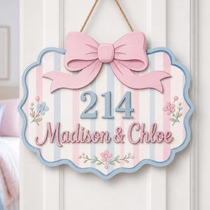 May include: A decorative wall sign featuring a light pink and blue striped background with a large pink bow at the top. The sign displays the number 214 and the names Madison & Chloe in blue and pink lettering, accented with small floral illustrations.