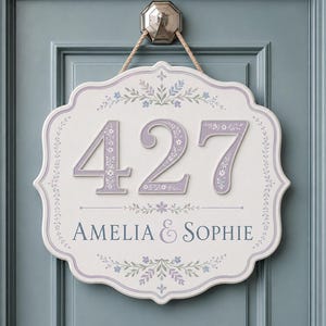 May include: A white, scalloped-edge wall plaque featuring the number 427 in large, light purple, floral-patterned digits. Below the number, the text AMELIA & SOPHIE is printed in dark blue, surrounded by delicate blue and green floral motifs.