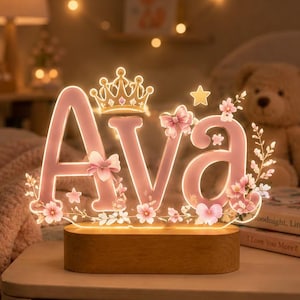 Könnte beinhalten: Ein personalisiertes LED-Lichtschild mit dem Namen Ava in rosa Buchstaben, montiert auf einem rechteckigen Holzsockel. Das Design zeigt eine goldene Krone, einen kleinen Stern, rosa Schleifen und florale Akzente mit warmem Licht.