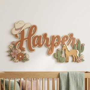 May include: A wooden wall sign featuring the name Harper in cursive script, decorated with a cowboy hat, flowers, a horse, and cacti in muted earth tones. Below is a light wood cot with a sage green blanket and several throw pillows.