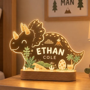 K&ouml;nnte beinhalten: Ein leuchtendes Nachtlicht in Triceratops-Form aus klarem Acryl mit Holzsockel. Der Dinosaurier zeigt aufgedruckte Illustrationen von Palmen, einem Vulkan und einem Ei sowie den personalisierten Text ETHAN COLE in zentrierten wei&szlig;en Buchstaben.
