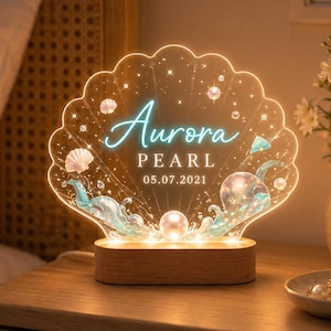 May include: A shell-shaped acrylic night light with a wooden base, featuring the name Aurora, the word PEARL, and the date 05.07.2021. The design includes illustrations of seashells, waves, and small pearls, illuminated by a warm internal light.