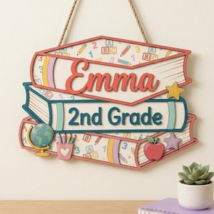 Puede incluir: Un letrero de madera colgante con forma de pila de tres libros y una cuerda. El libro superior muestra el nombre Emma en letra roja. El libro central presenta el texto 2nd Grade en azul. El diseño incluye un globo terráqueo, lápices y una manzana.