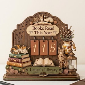 May include: A wooden decorative book tracker featuring the text "Books Read This Year" and "Laura's Library" with three hanging number tiles displaying 115. The piece includes carved cat figures, stacks of books, a vase with pussy willow, and a lantern.