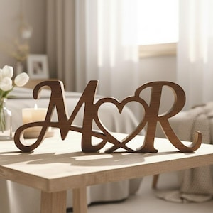 Puede incluir: Letrero decorativo de madera con las letras "M" y "R" unidas por un corazón. Las letras marrones oscuras están sobre una mesa de madera marrón claro. El fondo incluye un jarrón con flores blancas y una vela encendida. La mesa mide unos 60 cm de largo.