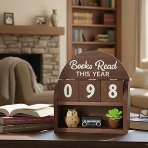 Może przedstawiać: Drewniana p&oacute;łka z napisem "Books Read THIS YEAR" w białym skrypcie. Na wyświetlaczu widnieje liczba "098" na trzech obrotowych klockach. Na p&oacute;łce znajduje się mała figurka sowy, miniaturowy boombox i doniczkowy sukulent.