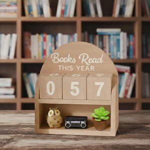 Może przedstawiać: Drewniany kalendarz blokowy z napisem "Books Read THIS YEAR" i liczbami "057". Kalendarz ma małą p&oacute;łkę z dekoracyjną sową, miniaturowym czarnym boomboxem i małym sukulentem w doniczce. Regały z książkami w tle.