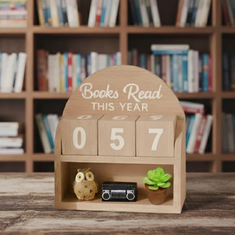 Może przedstawiać: Drewniany kalendarz blokowy z napisem "Books Read THIS YEAR" i liczbami "057". Kalendarz ma małą p&oacute;łkę z dekoracyjną sową, miniaturowym czarnym boomboxem i małym sukulentem w doniczce. Regały z książkami w tle.