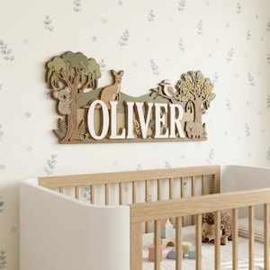 May include: A wooden wall sign with the name "OLIVER" in large, white letters. The sign features a koala, kangaroo, bird, and bear, along with trees and foliage. The sign is mounted above a crib with a wooden frame.