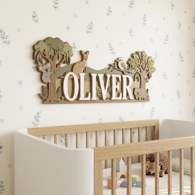 May include: A wooden wall sign with the name "OLIVER" in large, white letters. The sign features a koala, kangaroo, bird, and bear, along with trees and foliage. The sign is mounted above a crib with a wooden frame.