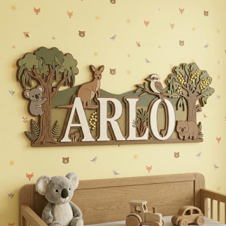 May include: A wooden wall hanging with the name "ARLO" in large white letters. The design features a koala, kangaroo, kookaburra, and wombat, along with trees and foliage, all in shades of brown, green, and cream. The background is a light yellow wall.
