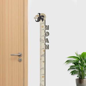 May include: A wooden height chart with the name "NOAH" in black letters. The chart features a whimsical design with a dragon and clouds at the top, and stars and clouds along the measuring scale. The scale is marked in inches, up to 180 inches.