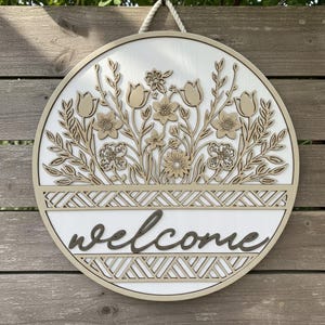 Pode incluir: Uma placa de boas-vindas redonda de madeira com fundo branco e design floral. A placa apresenta um arranjo floral cortado a laser em tons de bege e castanho, com a palavra "welcome" em fonte cursiva. Uma corda é anexada para pendurar.
