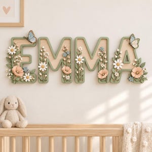 Könnte beinhalten: Ein hölzernes Wandschild mit dem Namen EMMA in salbeigrünen Buchstaben. Jeder Buchstabe ist mit dreidimensionalen weißen Gänseblümchen, rosa Blüten und kleinen blauen Blumen verziert. Zwei Schmetterlinge sind an den Buchstaben E und A befestigt.