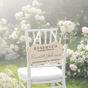 May include: A white chair with a sign that reads "RESERVED in loving memory of Scarlett Johanson GRANDMOTHER OF THE BRIDE." A bouquet of white roses and greenery is attached to the chair. The background features a garden with white flowers.