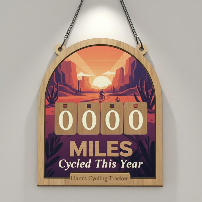 May include: Wooden cycling tracker with a desert landscape illustration. The design features a cyclist, mountains, and a setting sun. The tracker displays "0000 MILES Cycled This Year" and includes a space for a name. It hangs by a metal chain.