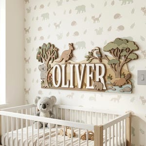 May include: Wooden name sign with the name "OLIVER" in white letters, featuring Australian animals like a kangaroo, koala, and platypus. The sign is set against a backdrop of a nursery with a crib and animal-themed wallpaper.