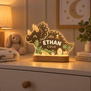 K&ouml;nnte beinhalten: Ein LED-Nachtlicht in Dinosaurierform mit Holzsockel, das eine Triceratops-Silhouette, Palmen, einen Vulkan und ein Ei zeigt. Die Lampe leuchtet und zeigt den personalisierten Text ETHAN COLE in wei&szlig;en Gro&szlig;buchstaben in der Mitte.