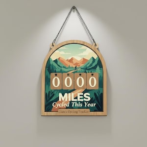 May include: Wooden cycling tracker with a mountain landscape design. The tracker displays "0000 Miles Cycled This Year" with a personalized name at the bottom. The frame is arched and hangs from a chain.