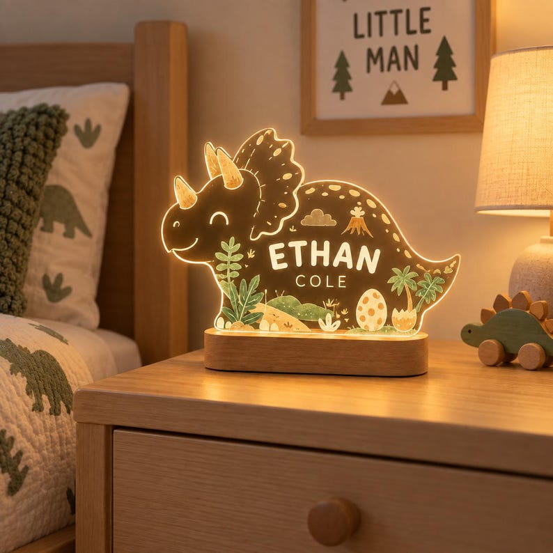K&ouml;nnte beinhalten: Ein leuchtendes Nachtlicht in Triceratops-Form aus klarem Acryl mit Holzsockel. Der Dinosaurier zeigt aufgedruckte Illustrationen von Palmen, einem Vulkan und einem Ei sowie den personalisierten Text ETHAN COLE in zentrierten wei&szlig;en Buchstaben.