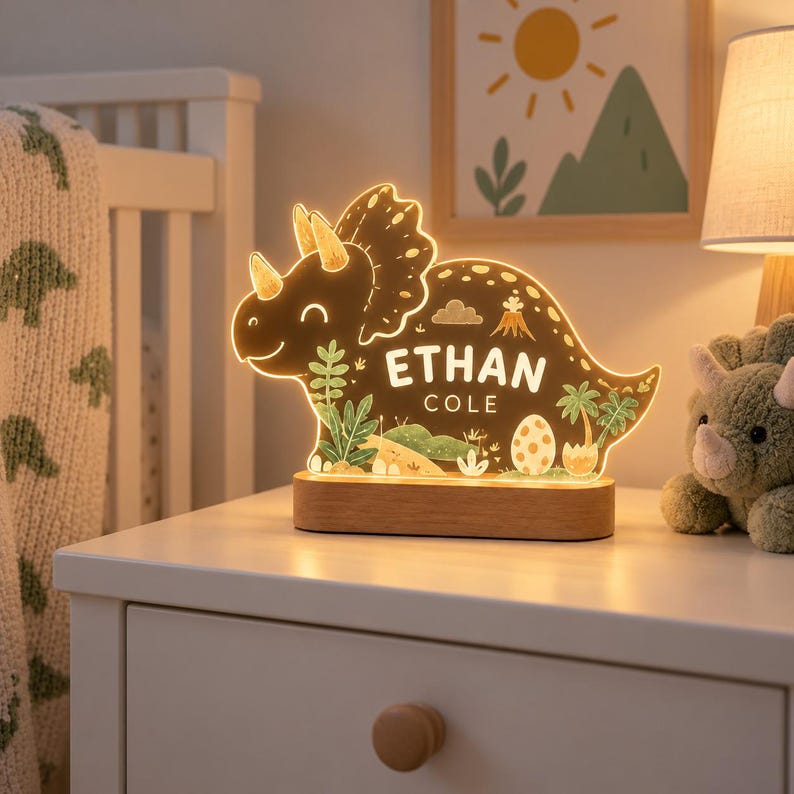 K&ouml;nnte beinhalten: Ein LED-Nachtlicht in Triceratops-Form mit warmem Lichtschein, auf dem der Name ETHAN COLE aufgedruckt ist. Das Design zeigt Illustrationen von Palmen, einem Vulkan und einem Dinosaurier-Ei, montiert auf einem rechteckigen Holzsockel.