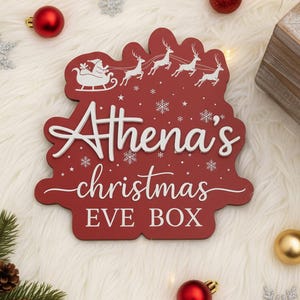 Könnte beinhalten: Rote Weihnachts-Eve-Box aus Holz mit dem Namen "Athena's" in weißer Schrift. Die Box zeigt ein Design von Santa's Schlitten und Rentieren sowie Schneeflocken. Dekorative Weihnachtskugeln umgeben die Box.