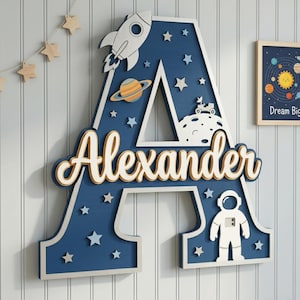 Könnte beinhalten: Ein großer, blauer Holzbuchstabe "A" mit dem Namen "Alexander" in Schreibschrift. Der Buchstabe ist mit einer Rakete, Planeten, Sternen und einem Astronauten verziert. Ein gerahmtes Bild mit Planeten ist sichtbar.