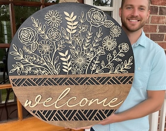 May include: A round wooden welcome sign with a floral design. The sign features laser-cut flowers and leaves in a light color against a black background. The word "welcome" is in a cursive font. The bottom section has a geometric pattern.