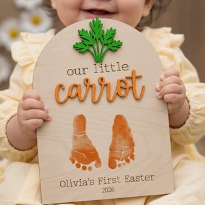 May include: Wooden plaque with the words "our little carrot" in orange script, topped with a green carrot top. Features two orange baby footprints and the text "Olivia's First Easter 2026".