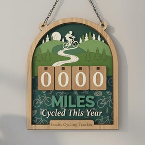 May include: Wooden cycling tracker with a green and white landscape design. Features a cyclist graphic, a winding road, and the text "MILES Cycled This Year." Includes a counter with the number "0000."