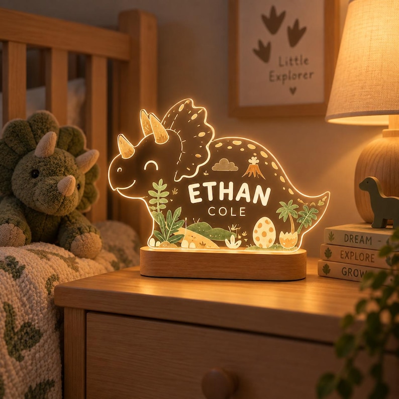 K&ouml;nnte beinhalten: Ein leuchtendes Nachtlicht aus Acryl in Triceratops-Form auf einem rechteckigen Holzsockel. Die Leuchte zeigt ein aufgedrucktes pr&auml;historisches Landschaftsmotiv mit Palmen, einem Vulkan und einem Ei sowie den Text ETHAN COLE in der Mitte.