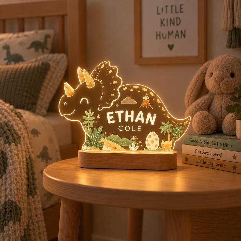 K&ouml;nnte beinhalten: Ein leuchtendes Nachtlicht in Triceratops-Form aus klarem Acryl mit einem Holzsockel. Der Dinosaurier zeigt aufgedruckte Illustrationen von Pflanzen, einem Vulkan und einem Ei sowie den personalisierten Text ETHAN COLE in wei&szlig;er Schrift.