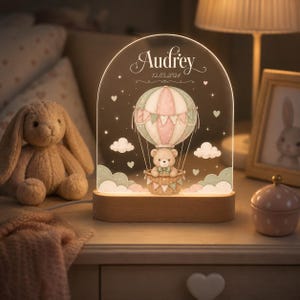 May include: An arched acrylic night light with a wooden base featuring a printed illustration of a teddy bear in a hot air balloon, the name Audrey, and the date 12.03.2024. The design includes pastel clouds, stars, and hearts with soft illumination.