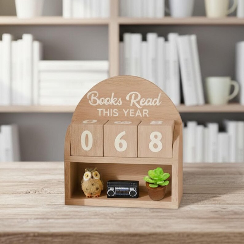 Może przedstawiać: Drewniany licznik książek z napisem "Books Read THIS YEAR" i liczbami "068". Zawiera małą figurkę sowy, miniaturowy boombox i doniczkę z sukulentem. Tło przedstawia p&oacute;łkę z książkami.