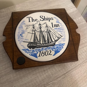 May include: A wooden coaster with a ceramic insert featuring a black and white illustration of a ship. The text "The Ships Inn" and the year "1802" are printed on the insert. The coaster has a dark brown wooden frame.