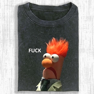 Peut inclure: T-shirt gris foncé avec un personnage de type Muppet aux cheveux orange, de grands yeux blancs et un nez rond orange. Le mot "FUCK" est imprimé au-dessus du personnage. Le t-shirt est plié.