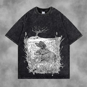 May include: Dark grey t-shirt with a distressed finish. The front displays a detailed black and white illustration of a rabbit in a natural setting, surrounded by plants and a tree. The design has a vintage, artistic aesthetic.