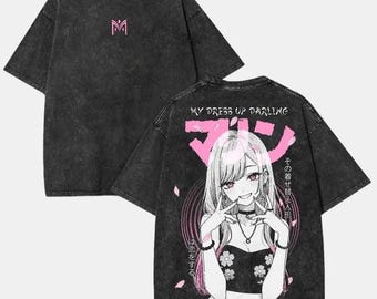 Puede incluir: Camiseta gris oscuro con un logotipo "M" rosa en la parte delantera. La parte trasera presenta un gráfico de un personaje de anime con el texto "MY DRESS UP DARLING" y texto japonés. El diseño incluye detalles florales y acentos rosas.