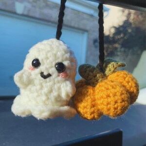 May include: Crocheted Halloween-themed car charm featuring a smiling white ghost with rosy cheeks and a cluster of orange pumpkins. The ghost has black eyes and a black mouth. The pumpkins have green stems.