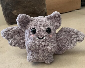 May include: A handmade, gray crochet bat with black safety eyes, small fangs, and blush on its cheeks. The bat has large, textured wings and ears. This plush toy is a cute, whimsical item.