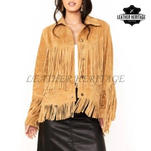 Può includere: Giacca in pelle scamosciata color cammello con frange sulle maniche, tasche sul petto e orlo. La giacca ha una chiusura con bottoni sul davanti e un colletto. Il testo "LEATHER HERITAGE" è stampato sulla giacca. Il logo Leather Heritage si trova nell'angolo in alto a destra.