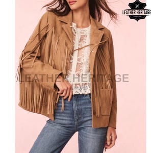 May include: A tan suede jacket with fringe detailing on the sleeves and chest. The jacket has a classic blazer cut with pockets and is paired with a white lace top and blue jeans. The "Leather Heritage" logo is visible.