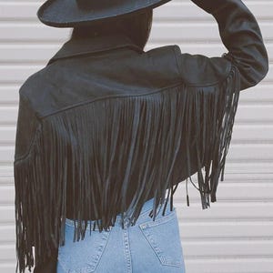 May include: Black suede jacket with long fringe detail across the back. The jacket has a classic collar and long sleeves. A black hat with a wide brim is worn with the jacket. Light blue denim jeans complete the look.