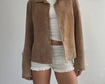 May include: A light brown suede jacket with a button-up front and a collared neckline. The jacket is worn over a white top and paired with white shorts. The jacket has long sleeves and a classic, tailored fit.