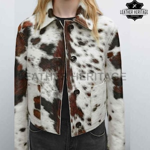 Könnte beinhalten: Eine kurze Jacke mit Kuhfellmuster in Weiß, Braun und Schwarz. Die Jacke hat einen Kragen, einen Knopfverschluss und zwei Vordertaschen. Das Markenlogo "LEATHER HERITAGE" ist oben rechts sichtbar.
